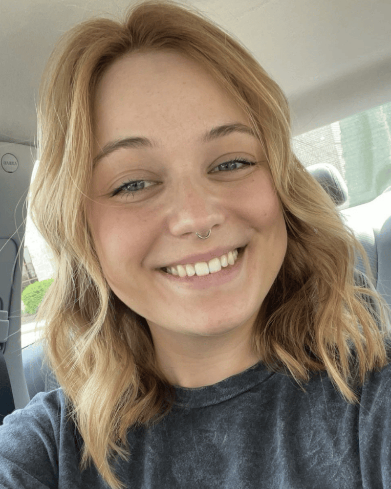 Close-up selfie of a smiling person with shoulder-length blonde hair and a septum piercing, seated in a car.